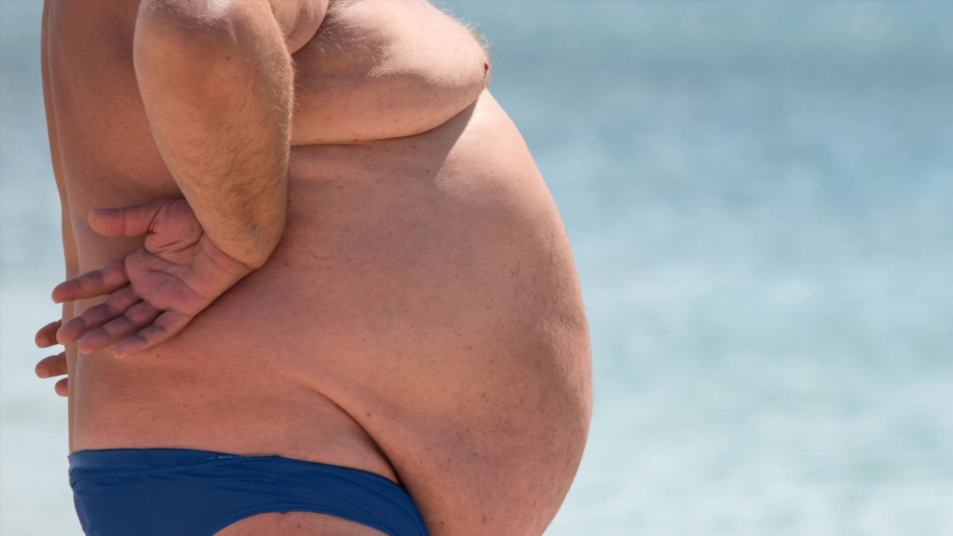 obese man on beach in small bathing suit with giant belly hanging in front